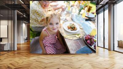 Portrait of a beautiful little girl sitting at table eating grilled food outdoors. Girl at family garden party. Wall mural