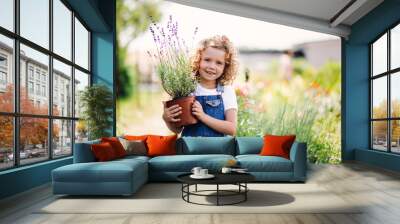 Portrait of small girl standing in the backyard garden, holding a plant in a pot. Wall mural