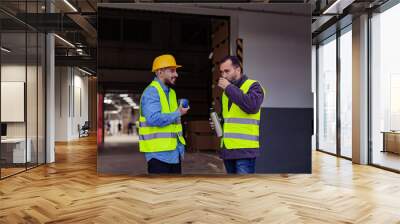 Portrait of two workers taking break from work, drinking coffee, talking and resting, standing on loading dock, outdoors. Colleagues drinking warm tea from thermos. Wall mural