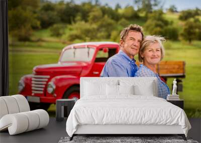 Senior couple hugging, vintage styled red car, sunny nature Wall mural