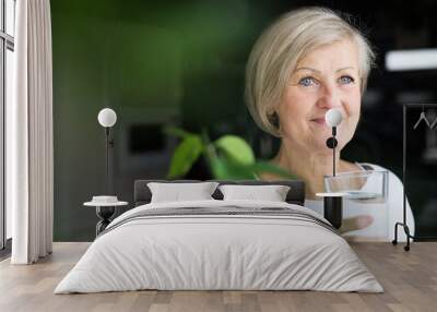 Senior woman holding a glass of water in the kitchen. Wall mural