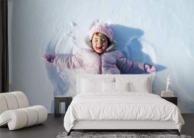 Top view portrait of cheerful small girl lying in snow in winter nature, making angels. Wall mural