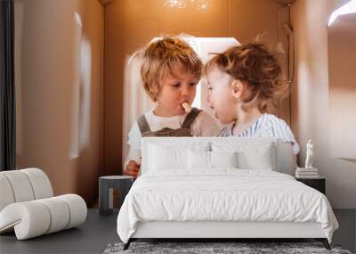 Two toddler children playing indoors in cardboard house at home, eating. Wall mural