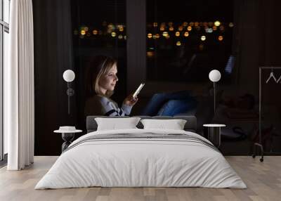 Woman at desk, holding smartphone, working on laptop at night. Wall mural