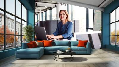 Young businesswoman with laptop sitting in an office, looking at camera. Wall mural