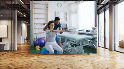 Young male physiotherapist exercising with young woman patient on ball in a physic room Wall mural