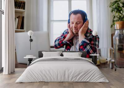 Young man with Down syndrome sitting at desk in office and using laptop, listening to music from headphones. Wall mural