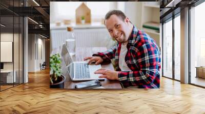 Young man with Down syndrome sitting at desk in office and using laptop, looking at camera and smiling. Wall mural