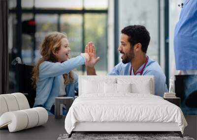 Young multiracial doctor having fun with little girl on wheelchair. Wall mural