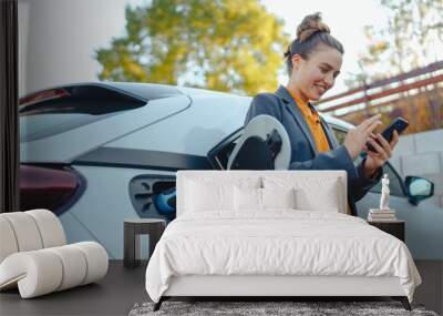 young woman with smartphone waiting while her electric car charging in home charging station, sustai Wall mural