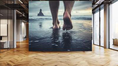 Male adult walking barefoot on a dark gray sand beach, with waves pounding under his feet or legs. Wall mural