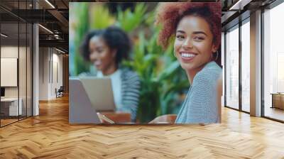 There are two professional women in an office space that's well lit, has hanging plants, and has large windows. One wears an olive green blouse with beige trousers, and leans forward. Wall mural