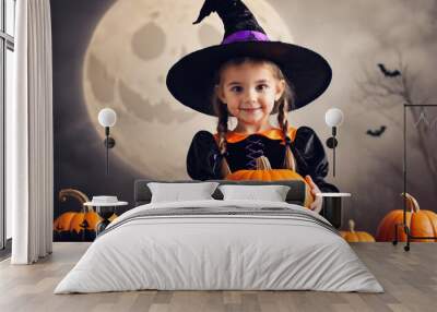 Young girl in a witch costume holding a pumpkin near carved decorations Wall mural