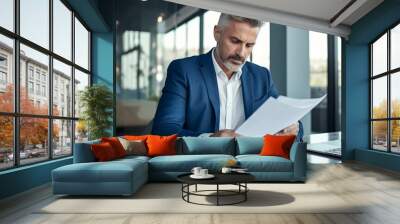 A business man in a suit sitting at his desk and reading documents while using a laptop computer in an office background with glass windows. Wall mural