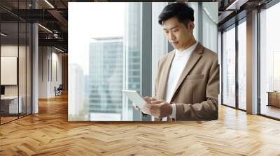 A handsome Asian man in casual attire stands by the window, holding an iPad and looking at it intently as he relaxes after work.  Wall mural