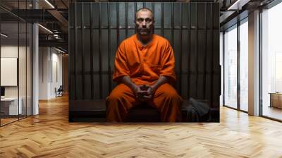 A man dressed in orange sits on a bench of a prison cell alone, looking at the camera, jail or imprisonment concept image. Wall mural