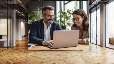 A middle-aged man and woman in casual attire are sitting at an office desk with a laptop computer open on the table, facing the camera and smiling while talking to each other.  Wall mural