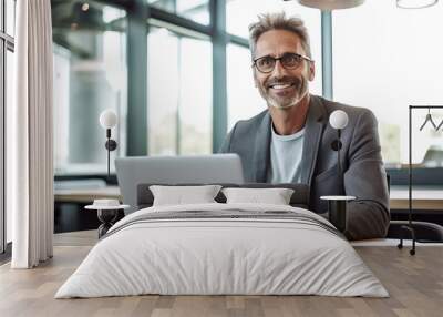 A photo of a smiling middle-aged man in business casual attire sitting at an office desk, working on his laptop. Wall mural