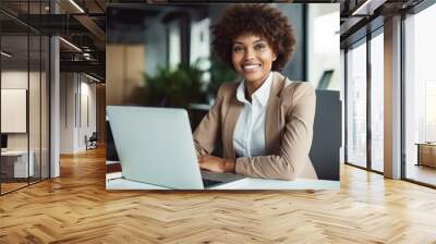 A smiling Black businesswoman sitting at her desk in an office, working on a laptop computer. Wall mural