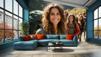 Portrait of a happy smiling pretty brunette girl waving hand looking cheerful at camera while trekking with backpack during a walking tour of the mountains with a group of her young friends. Wall mural