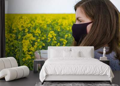 Girl in a mask on her face holding coffee among a flowering field of rapeseed in cloudy weather
 Wall mural