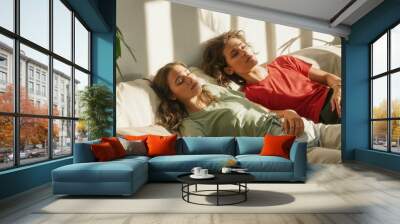 Two people lying on couches in an open space with white walls and green plants. The woman is wearing a red tshirt while the man wears a light grey long-sleeved shirt. They both have their eyes closed Wall mural