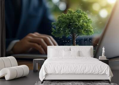 A businessman's hands type on a laptop with a tree growing out of the screen, symbolizing sustainable growth. Wall mural