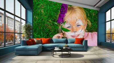 beautiful little girl blonde on a children's holiday with a pattern of a lion cub on her face, emotionally posing while lying on bright green grass holding a flower in her teeth Wall mural