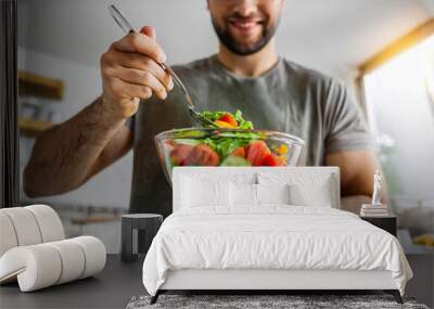 Young man eating a healthy vegan salad from a glass bowl in the kitchen. Vegan food concept Wall mural