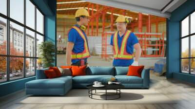 Two construction workers in safety gear talking inside a modern industrial warehouse with red steel framework and various construction materials. Wall mural