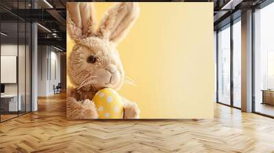 A close-up of a bunny plush toy holding an Easter egg on a light yellow background Wall mural