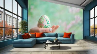 A close-up of a hand holding a beautifully decorated Easter egg on a soft green background Wall mural