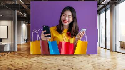 Happy Asian woman with shopping bags and phone, enjoying online shopping on purple background, e-commerce concept. Wall mural