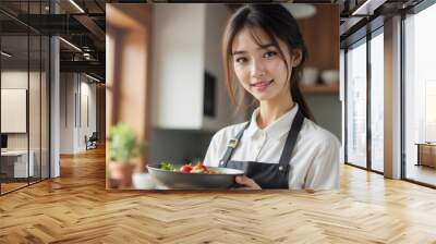 A cheerful woman in an apron holds a fresh salad in her kitchen, showcasing her culinary skills and passion for healthy cooking. Wall mural