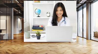 Asian female doctor sitting at hospital office desk giving all patient convenience online service advice and smiling write a prescription on laptop to order medical,health care and preventing disease Wall mural