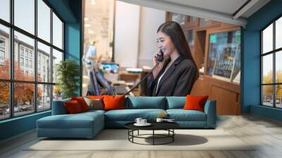 asian woman hotel receptionist holding a wired landline phone against her ear smiling looking down at a computer monitor with a keyboard and a mouse reading information on her desk behind the counter. Wall mural