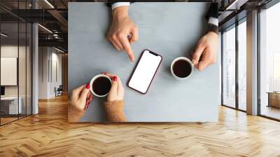 Business man in cafe showing blank phone screen to business woman with coffee cup on gray wooden table background. Top view. Wall mural