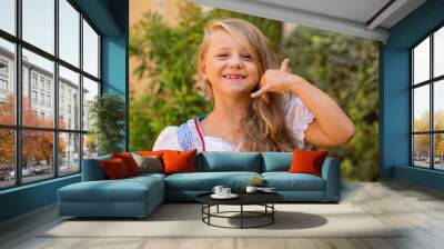 Call me sign. Cute little blonde girl child shows by gesture that she speaks on the phone smiling toothless isolated green shrubs nature garden near house outdoors on the background Wall mural