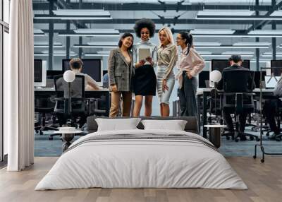 A group of young successful business women working together standing in the middle of the office and looking at a tablet. In the background are colleagues working on computers. Wall mural