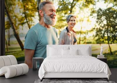 Active hobby on retirement. Grey haired caucasian man with beautiful wife strolling at summer park and smiling cheerfully. Fitness retired couple in sport attire taking break after morning run. Wall mural