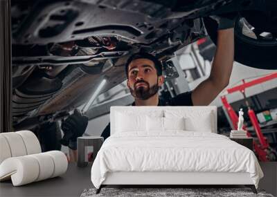 Auto mechanic in his workshop looking under a car on a hoist. Wall mural