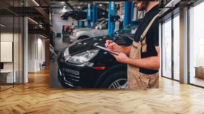 Cropped image of handsome mechanic in uniform making notes while standing in auto service. Wall mural