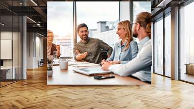 Four business people sitting along table while communicating and laughing in office space.  Businesswomen and businessmen in formal outfits exchanging professional experience indoors. Copy space. Wall mural