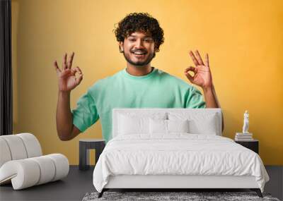 Handsome Indian man showing ok gesture, looking at camera and smiling while standing against yellow background. Wall mural