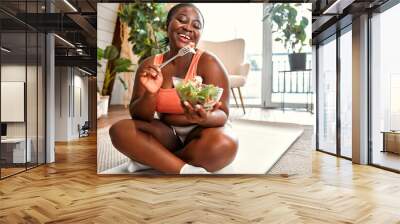 Healthy diet and sport. Beautiful plump woman in sport clothes eating vegetable salad from glass bowl while sitting on floor. Black young female following slimming and exercising program at home. Wall mural