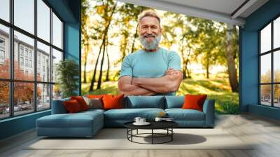 Healthy retirement lifestyle. Muscular grey bearded man keeping arms crossed and smiling while posing at green city park. Caucasian pensioner in sport clothes feeling happiness from active hobby. Wall mural