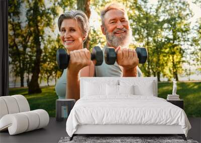 Joint workout on retirement. Beautiful and healthy elderly couple keeping dumbbells in hands while standing back to back at sunny park. Smiling family of two doing exercises for arms on fresh air. Wall mural