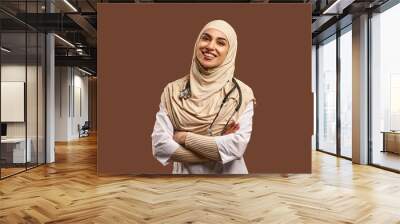 Portrait of a female Muslim doctor wearing a white coat, arms crossed, standing against a brown background and smiling. The concept of medicine, care, health. Wall mural