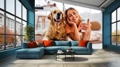 Saving memories with pet. Smiling woman with blond hair snuggling to furry friend and taking selfie on modern cell phone. Obedient golden retriever lying on floor near delighted female owner. Wall mural