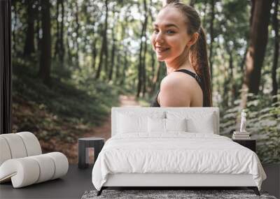 Smiling athletic woman before her run outdoors. Portrait of a young sporty woman smiling before training in the forest. Beautiful girl looking and smiling at the camera before run Wall mural